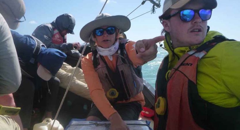 a person sitting on a sailboat points toward a spot in the distance while another looks on 